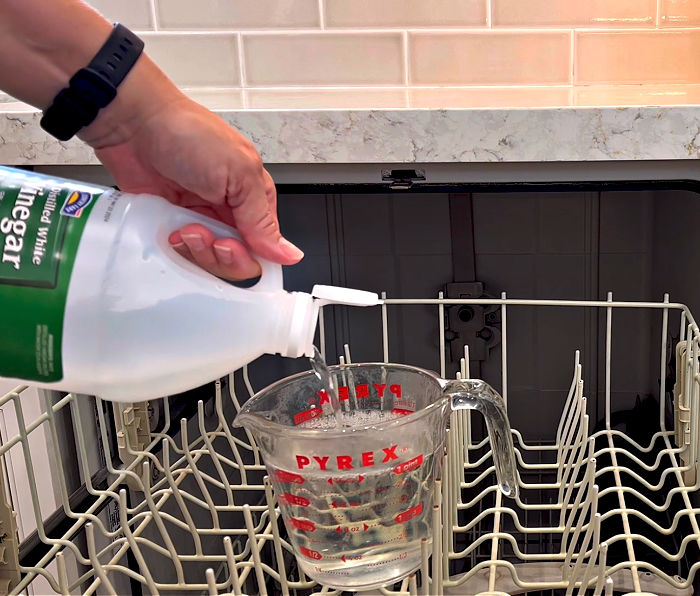 a person pouring white vinegar into a measuring cup inside an empty dishwasher