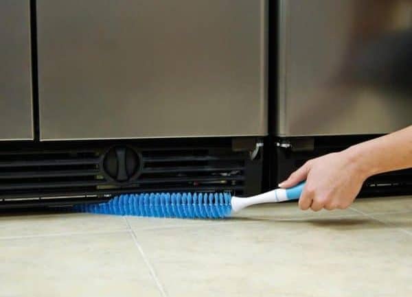 person using a refrigerator coil cleaning brush to remove dust and debris from the condenser coils located at the bottom of the refrigerator