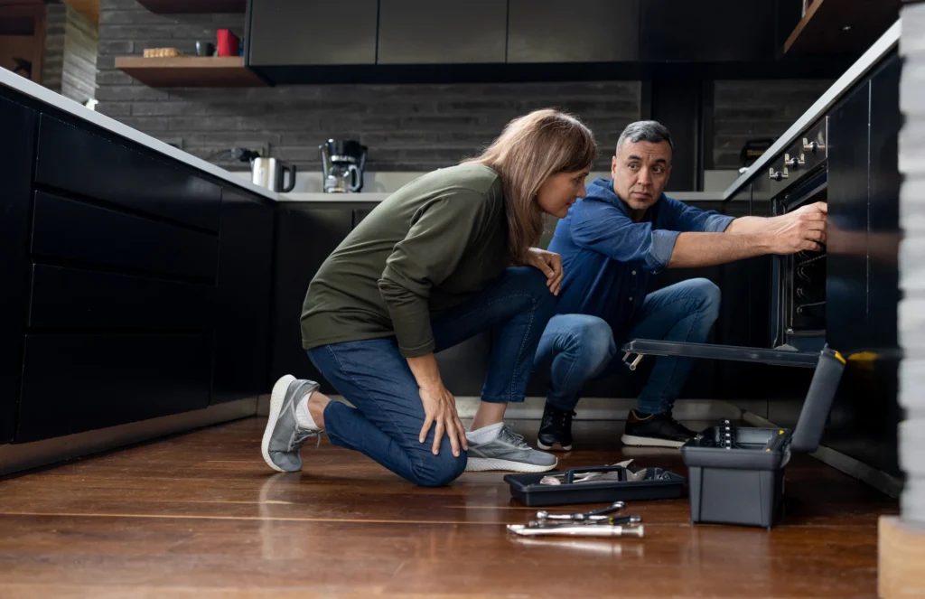 shows two people, possibly a couple or a homeowner and a technician, inspecting an appliance, likely an oven or stove, in a modern kitchen. 