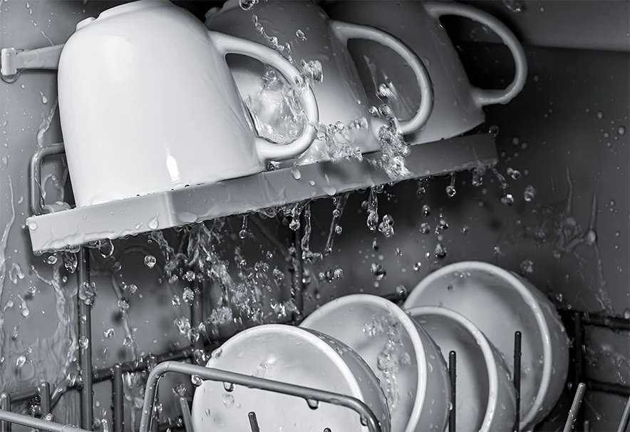 the interior of a dishwasher in operation, with water spraying onto dishes and flowing down from the top rack onto the lower items. 