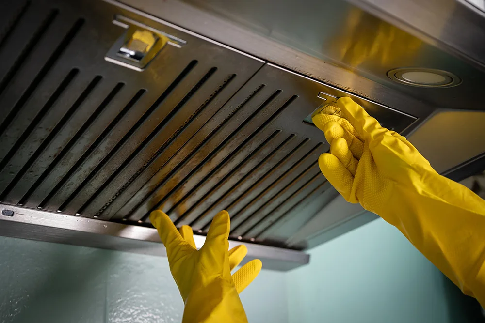 a person wearing yellow protective gloves attempting to remove a dirty filter from a stainless steel kitchen exhaust hood