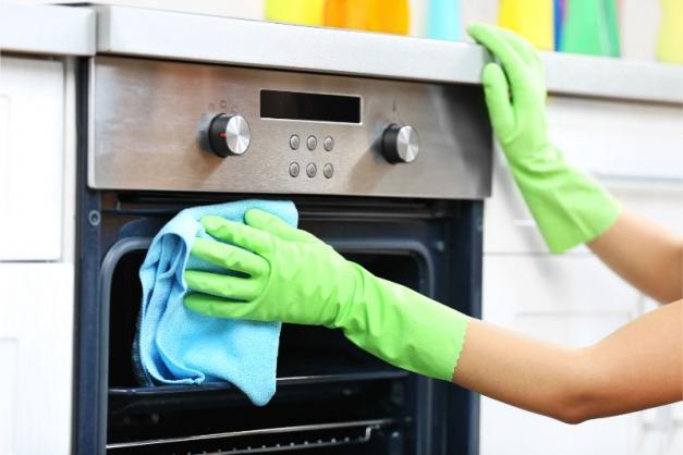 a person wearing green gloves and cleaning the interior of a stainless steel oven with a blue cloth